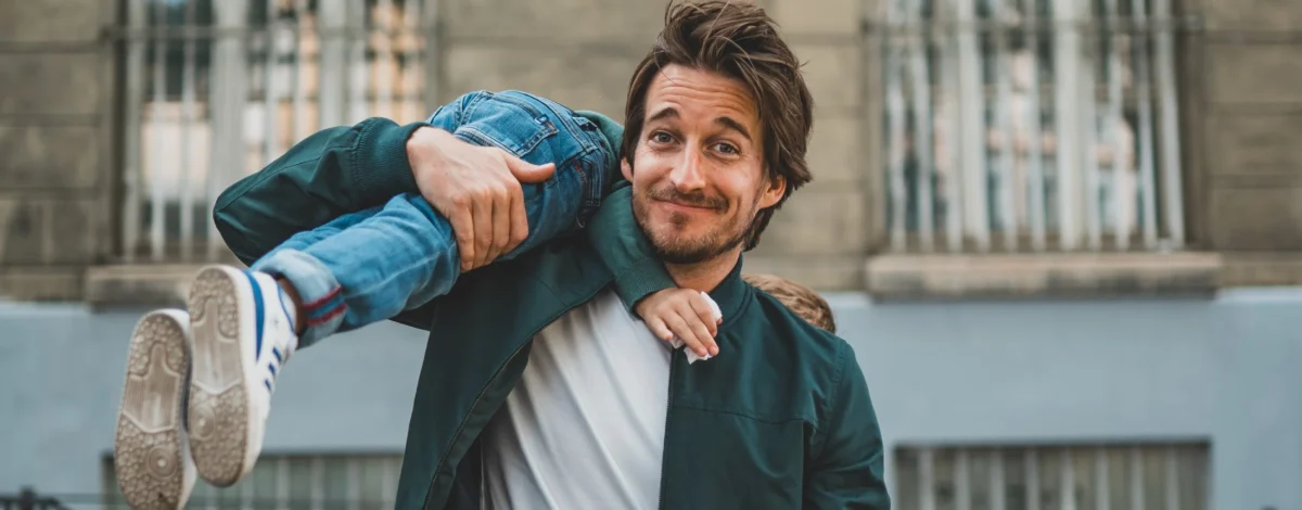 Un homme est assis et regarde la caméra, avec un enfant sur son épaule, en souriant.