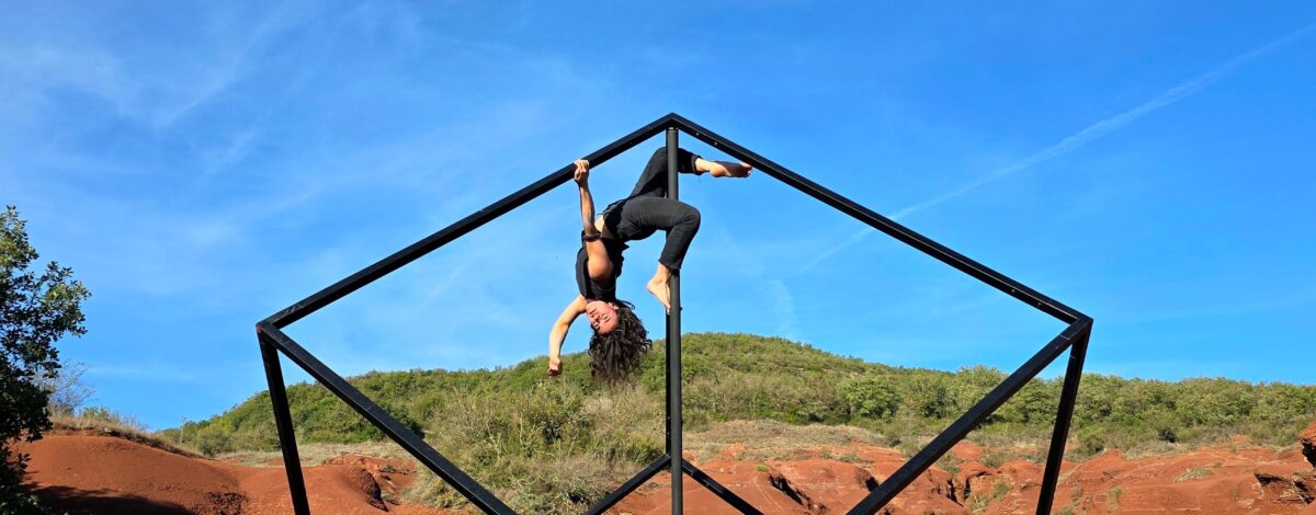 Une structure en métal, sorte de cube géant, est au centre de l'image. Une femme y est suspendue, par les jambes, comme en cochon pendu.