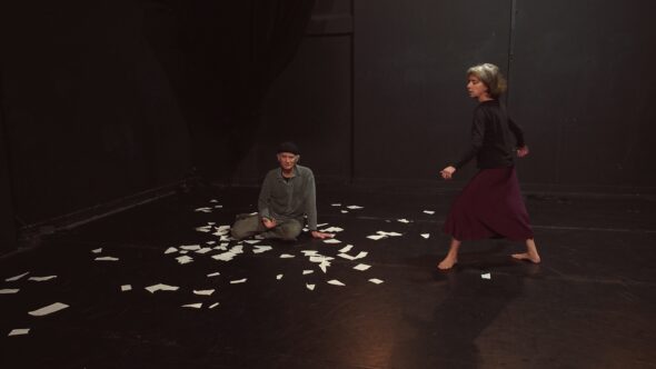 un homme âgé est assis par terre, des bouts de papier blanc déchirés sont disséminés autour de lui. Une femme est debout à sa gauche et semble être en train de danser